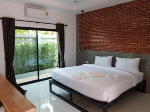 A hotel room with a bed, a wooden bench, a brick accent wall, and a view of greenery through a sliding door.