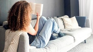 Una persona leyendo un libro en un sofá gris en una habitación con paredes oscuras y un espejo grande.