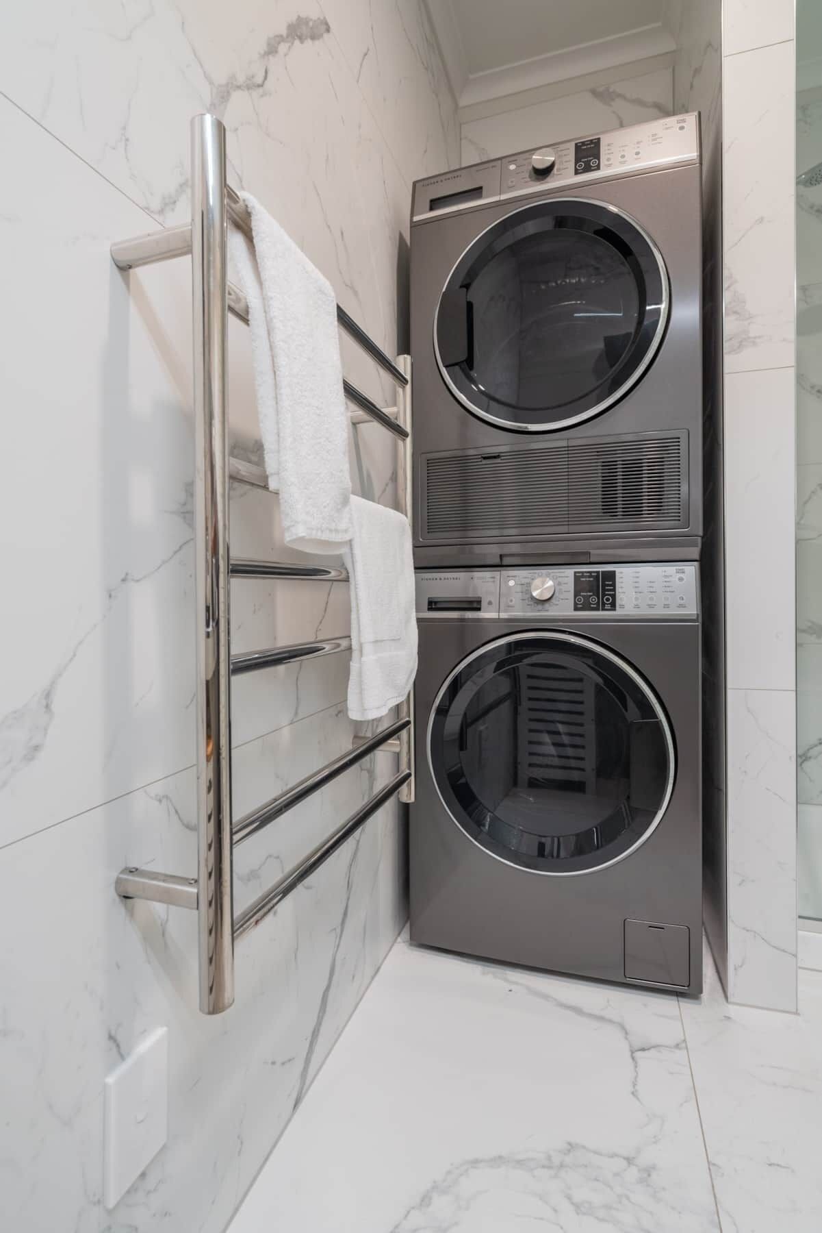 A compact, stainless steel wall-mounted washer and dryer unit in a bathroom with white marble tiles.