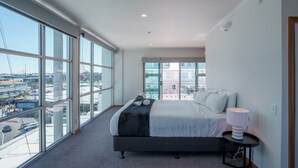 Une chambre d’hôtel moderne avec un grand lit, une table de chevet avec une lampe et une vue sur la ville grâce à de grandes fenêtres du sol au plafond.