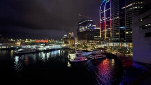 Une vue nocturne d’une ville, avec des bâtiments illuminés, un port de plaisance rempli de bateaux et un pont en arrière-plan.