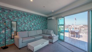 Un salon moderne avec un canapé d’angle gris, une table basse blanche et une grande fenêtre donnant sur un port de plaisance.