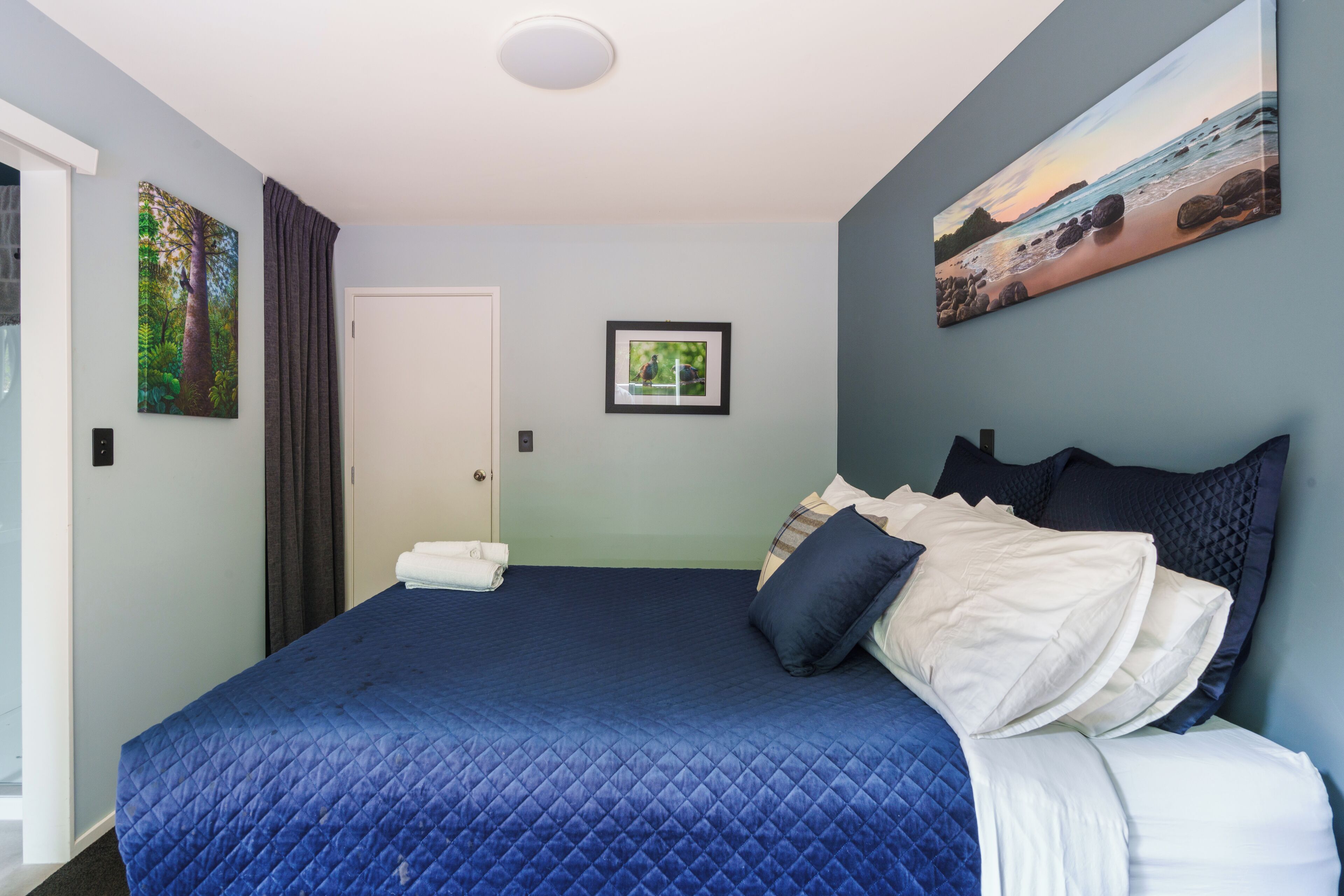 A neatly made bed with a blue quilt, a framed picture on the wall, and a door with a towel on the floor.