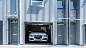 Un edificio de varios pisos con entrada para coches, un coche blanco estacionado en su interior y un cielo despejado.