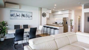 Ein modernes Wohnzimmer mit einem weißen Sofa, einem Essbereich mit Tisch und Stühlen, einer Küche mit weißen Schränken und einem Stadtbild an der Wand.