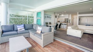 Ein modernes Wohnzimmer mit einem grauen Ecksofa, einem weißen Couchtisch und einem weißen Sessel. Es gibt eine Essecke mit Tisch und Stühlen, und durch eine offene Tür ist eine Küche erkennbar.