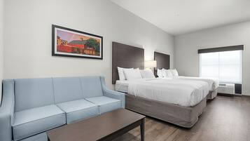 Habitación de hotel con un sofá azul claro, una mesa de centro de madera, dos camas individuales con sábanas blancas, un cuadro en la pared y un amplio ventanal que proporciona luz natural.