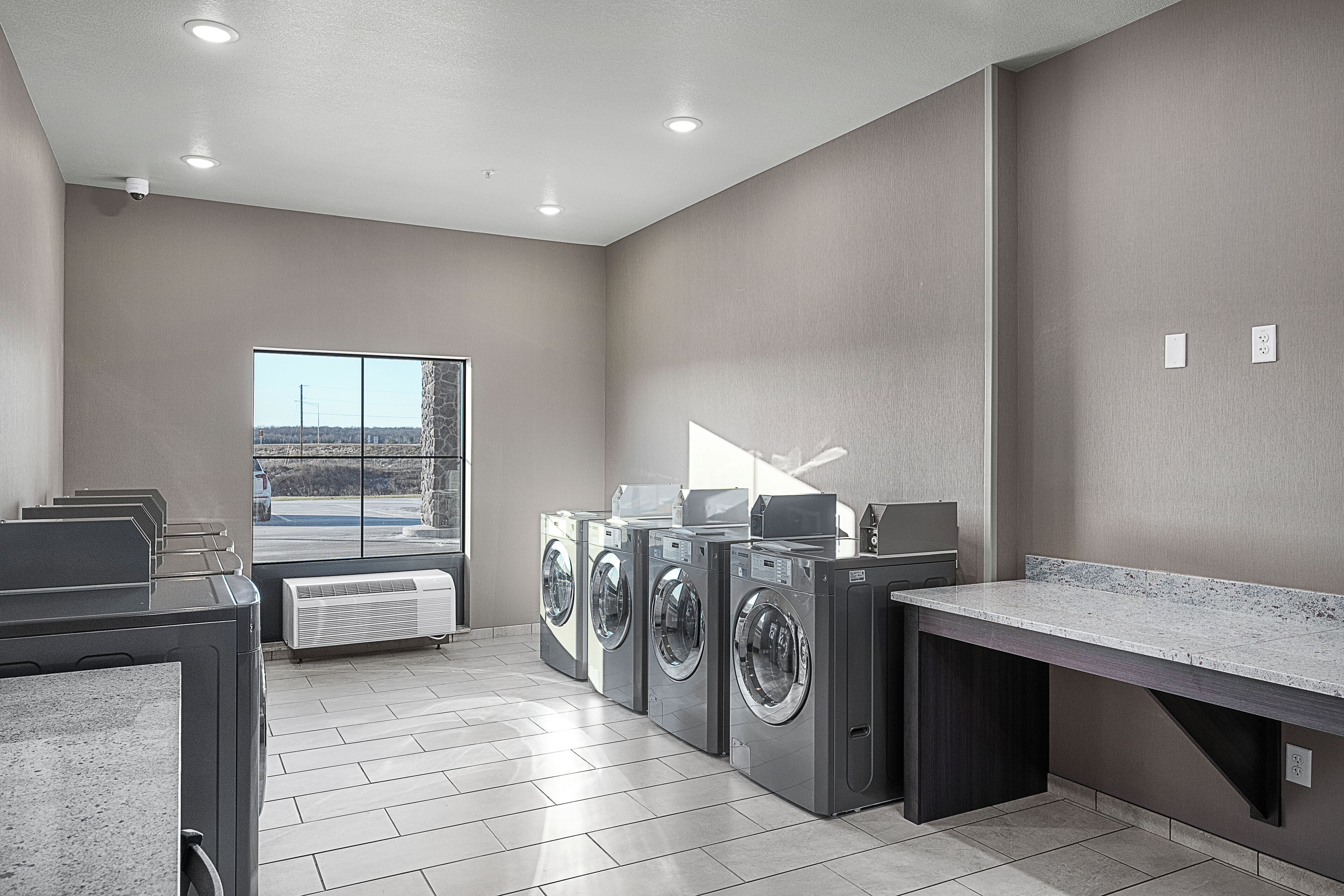 A laundry room with four washing machines, a large window, and a countertop.