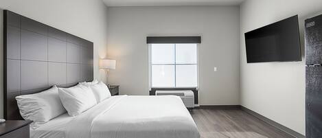 A modern hotel room with a large bed, a flat-screen TV, a window with blinds, and a built-in air conditioning unit.