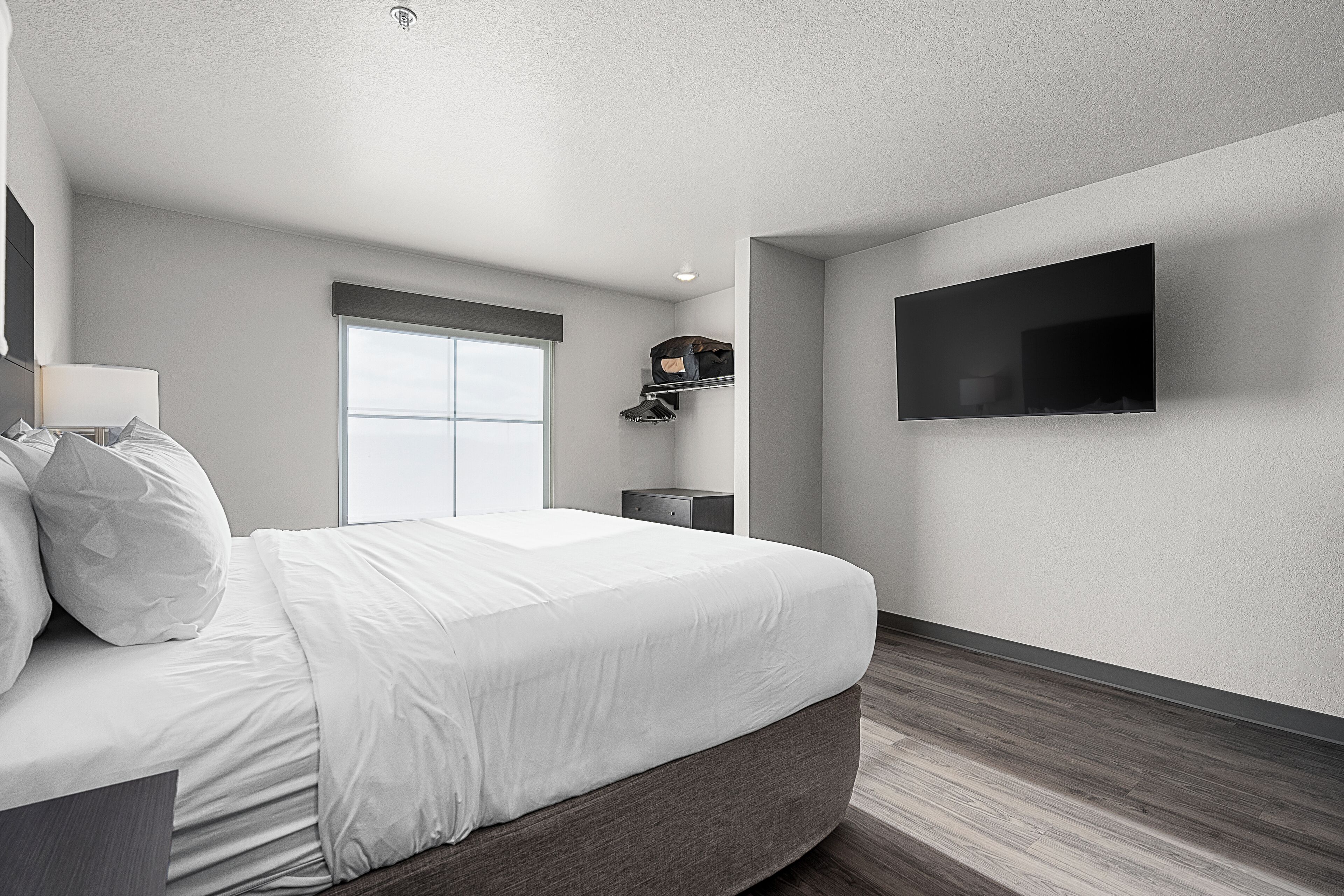 A modern hotel room with a large bed, a flat-screen TV mounted on the wall, a window with a view, and a small bedside table.