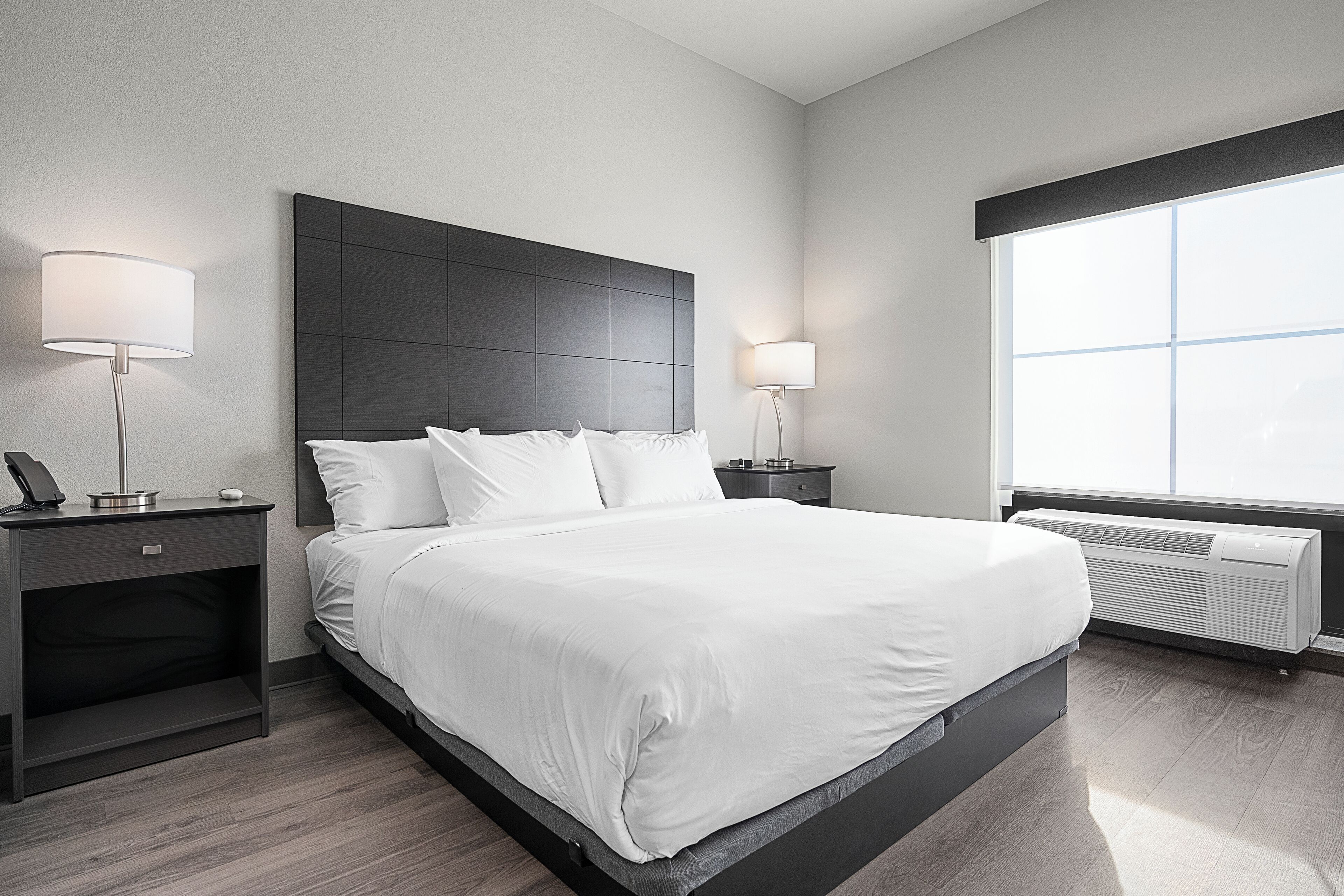 A modern hotel room with a large bed, two bedside tables with lamps, a window with blinds, and a wall-mounted air conditioner.
