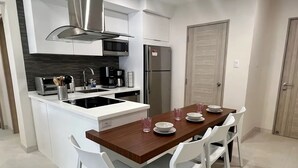 A modern kitchen with white cabinetry, a central island with a stovetop, a built-in oven, and a refrigerator. There is a dining table with white chairs and a few dishes and glasses on it. - THE Aracari (Noord)