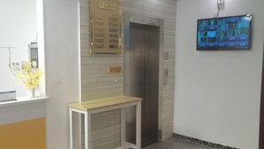 Zona de recepción con un mostrador de madera, un ascensor, un televisor montado en la pared y un tablón de anuncios con texto.