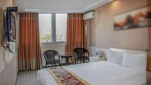Habitación de hotel con cama, escritorio, dos sillas, ventana con cortinas, equipo de aire acondicionado y un cuadro en la pared.