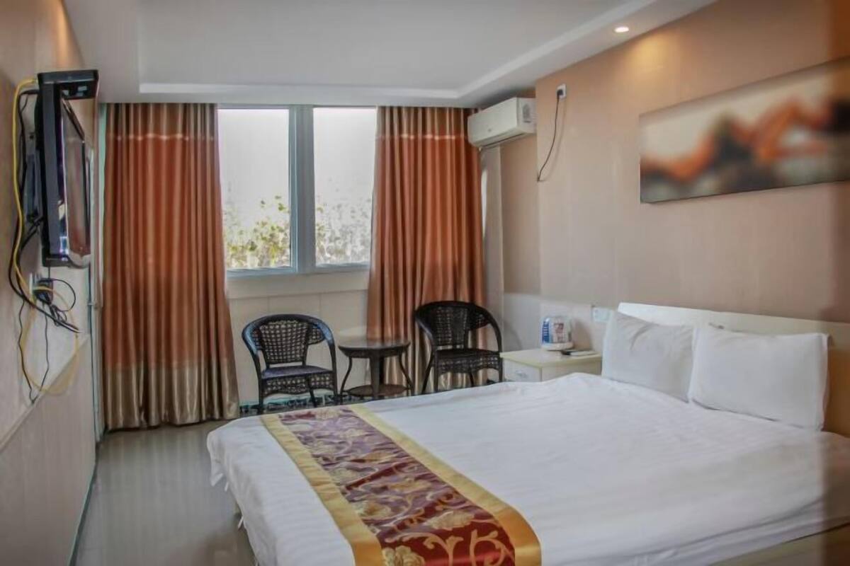 A hotel room with a bed, a desk, two chairs, a window with curtains, an air conditioning unit, and a painting on the wall.