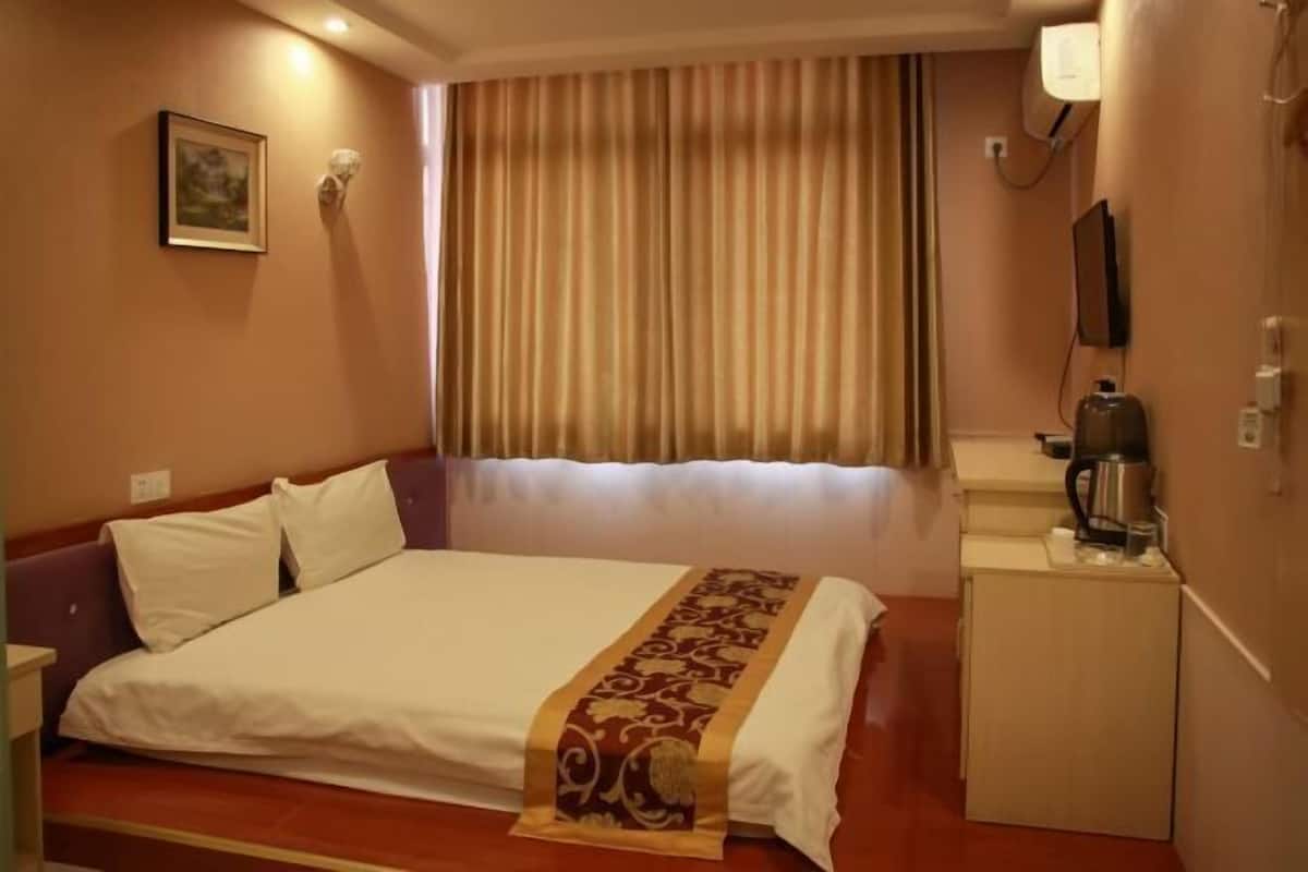 A hotel room with a bed, a small desk with a kettle, a television, and a window with curtains.