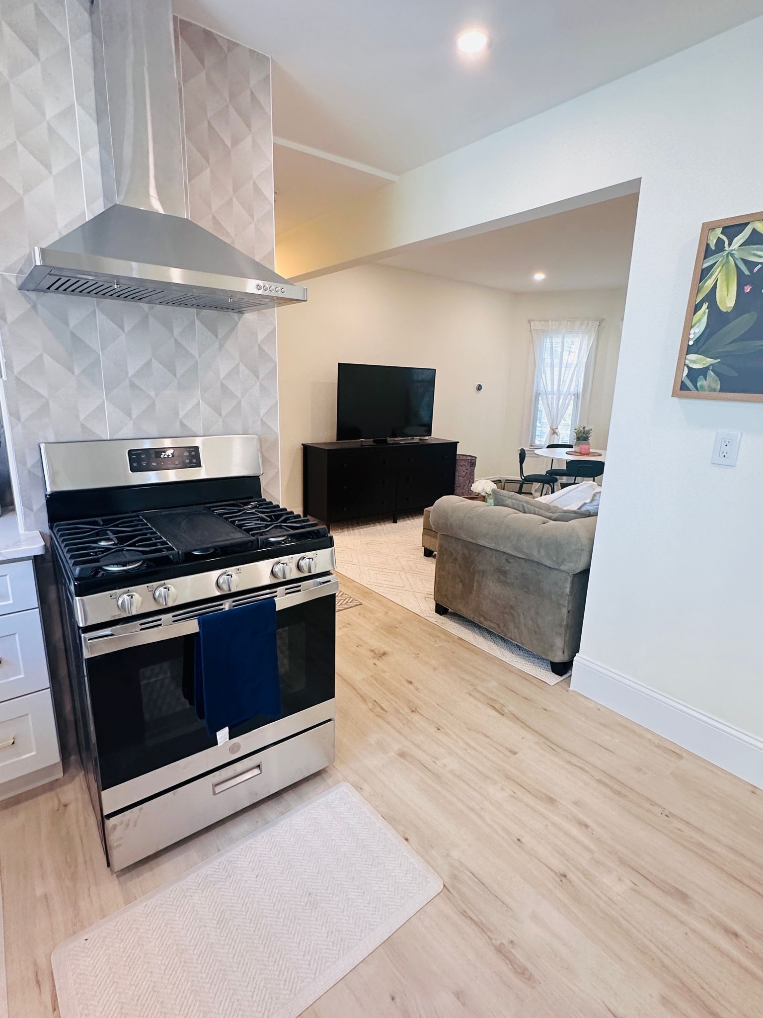 A modern kitchen with a stainless steel stove, a built-in oven, and a range hood. There is a dining area with a table and chairs, and a living room with a sofa and a television.