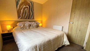 Ein ordentlich bezogenes Bett mit weißer Decke und zwei Kissen, einer Nachttischlampe und einem an der Wand hängenden abstrakten Gemälde.