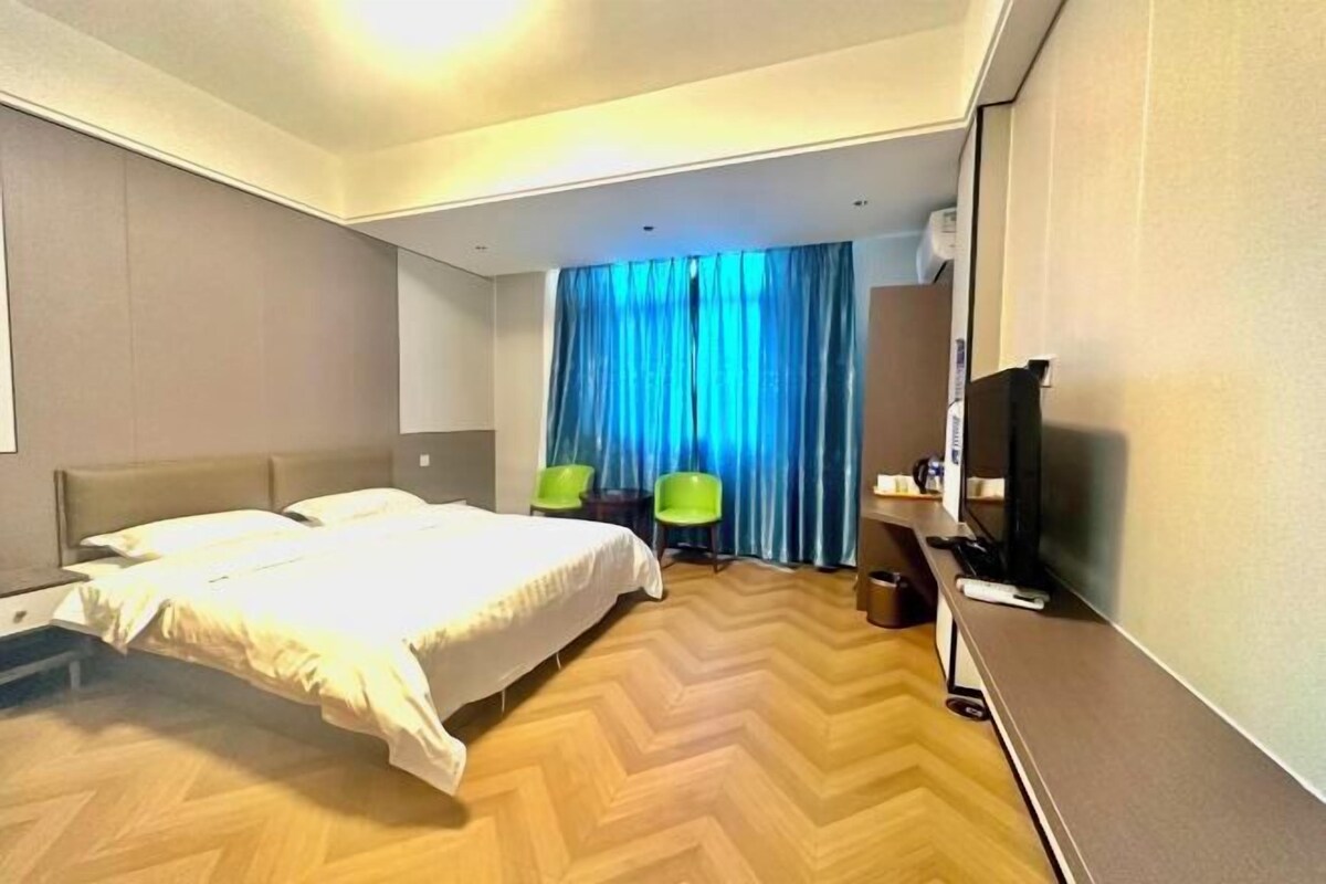 Habitación de hotel con cama, dos sillas, escritorio con televisor y un ventanal con cortinas.