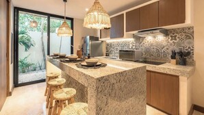 A modern kitchen with a central island, bar stools, hanging pendant lights, and a view of greenery outside. - Gem in the Jungle Private Pool Beach Club Access (Tulum)