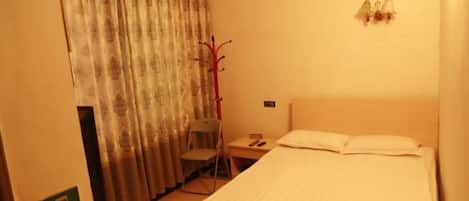 A hotel room with a bed, a chair, a desk, a curtain, and a wall-mounted light fixture.
