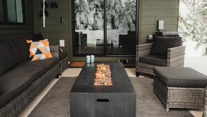 Un espace extérieur moderne, aménagé pour se détendre, avec un foyer, des meubles en osier et une vue sur les arbres enneigés, visible à travers une porte coulissante.