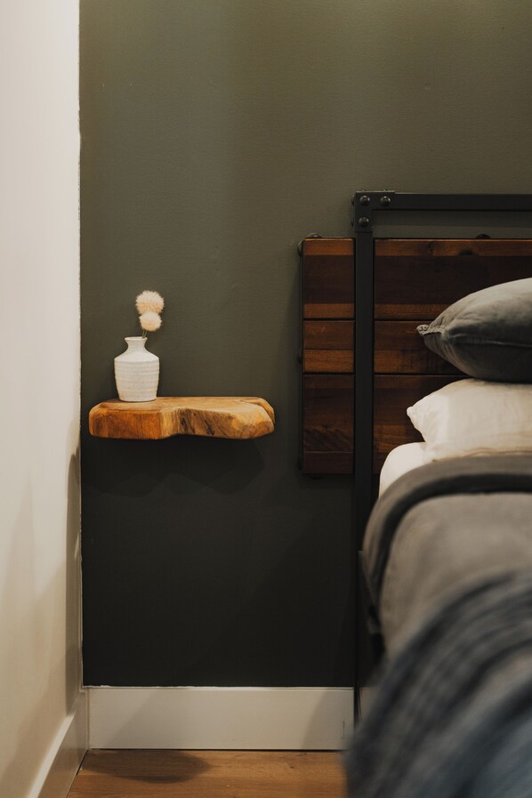 A bedroom with a dark wall, a wooden bedside table with a white vase, and a bed with a gray pillow. - Cedar Chalet: Ski-In/Ski-Out Suite with European Sauna, Firepit & King Bed. (Snoqualmie Pass)