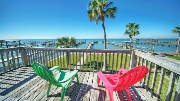 A wooden deck with two chairs, a palm tree, and a view of the ocean. - Fish - Relax - Repeat! Waterfront Retreat with Private Pier in Rockport, TX (Rockport)