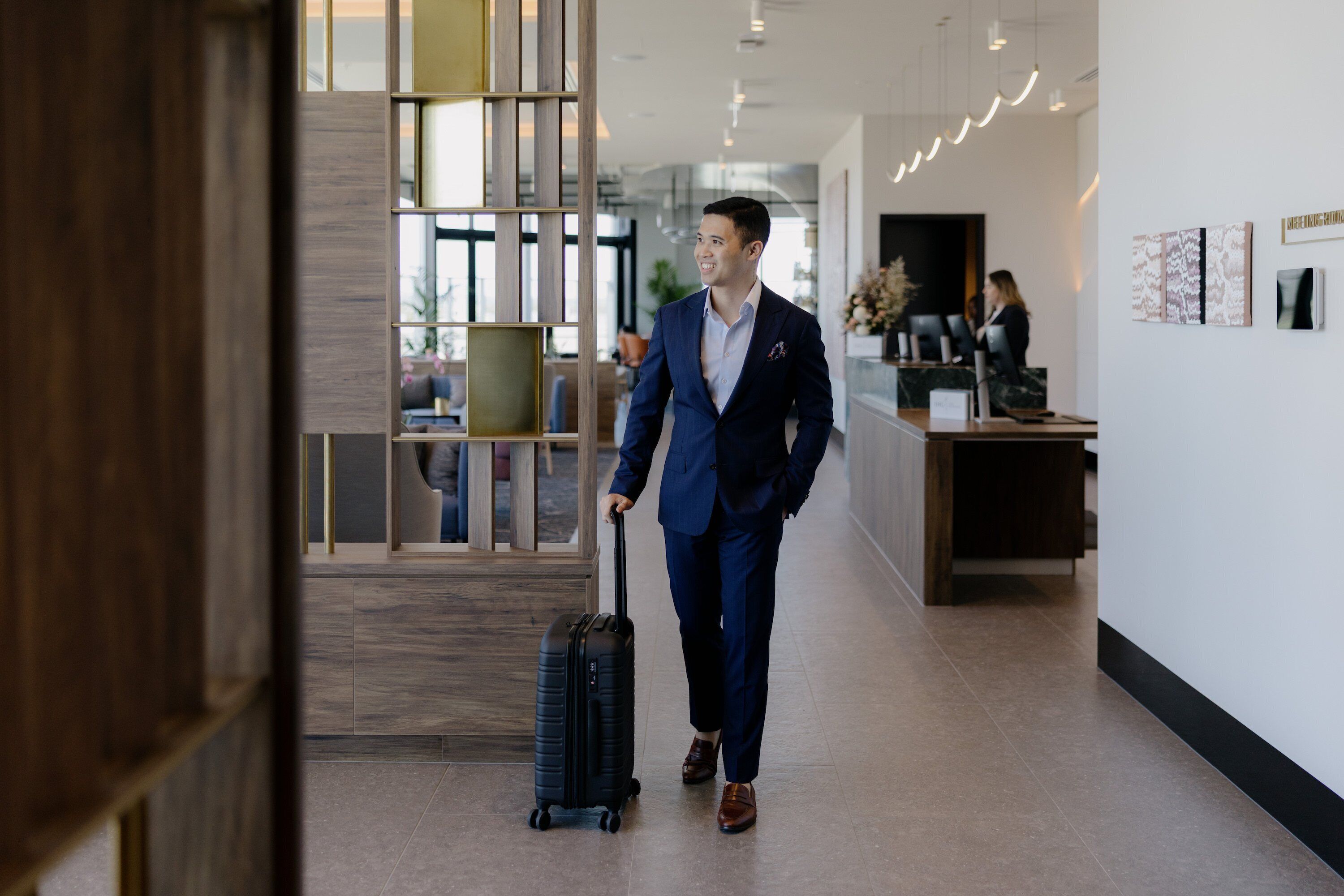 A man in a blue suit with a black suitcase walking through a modern hotel lobby with a reception desk and artwork on the walls.