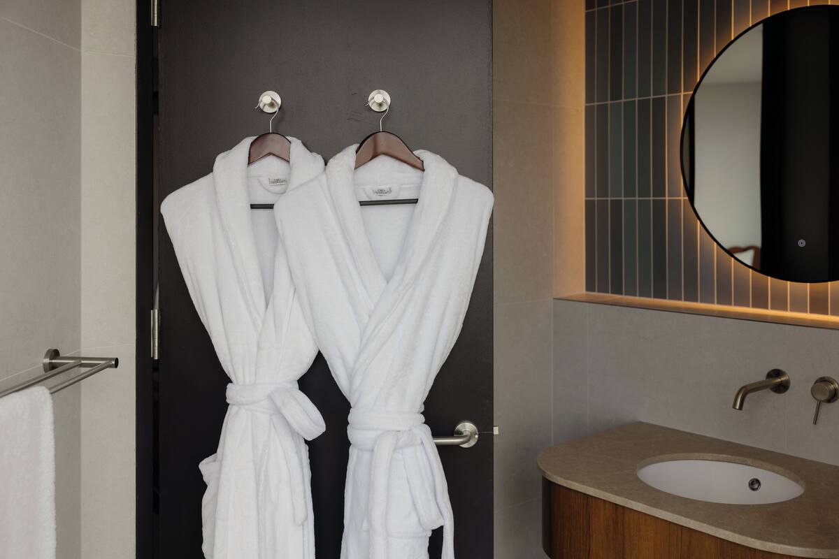 Ein Badezimmer mit zwei weißen Bademänteln, die an einer Tür hängen, einem runden Spiegel und einem Waschbecken mit einer Holztischplatte.