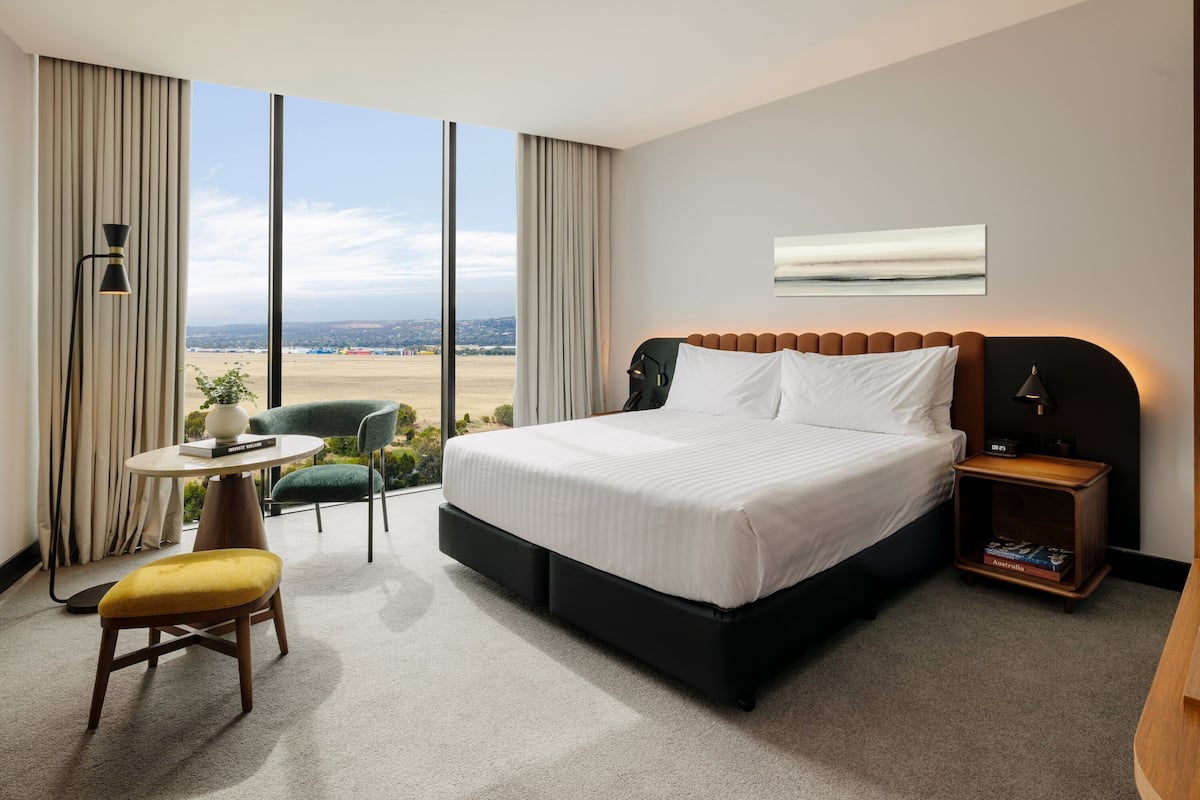 Ein Hotelzimmer mit einem großen Bett, einem kleinen Tisch mit einer Lampe, einem Sessel und Blick ins Grüne.