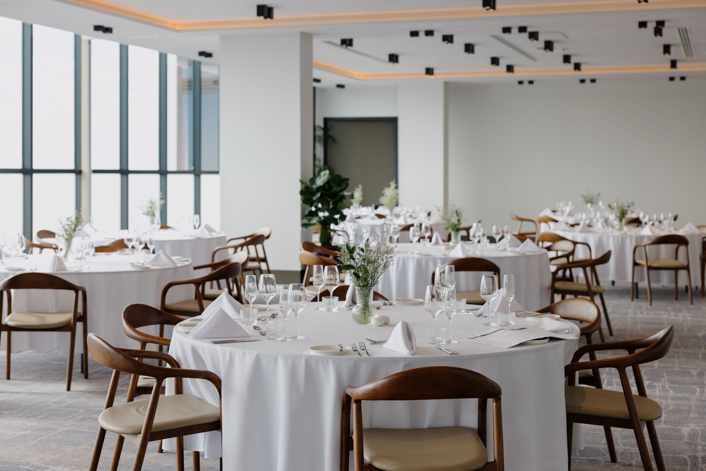 A dining area with round tables set for a formal event, featuring white tablecloths, wooden chairs, and floral centerpieces.