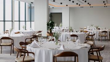 A dining area with round tables set for a formal event, featuring white tablecloths, wooden chairs, and floral centerpieces.