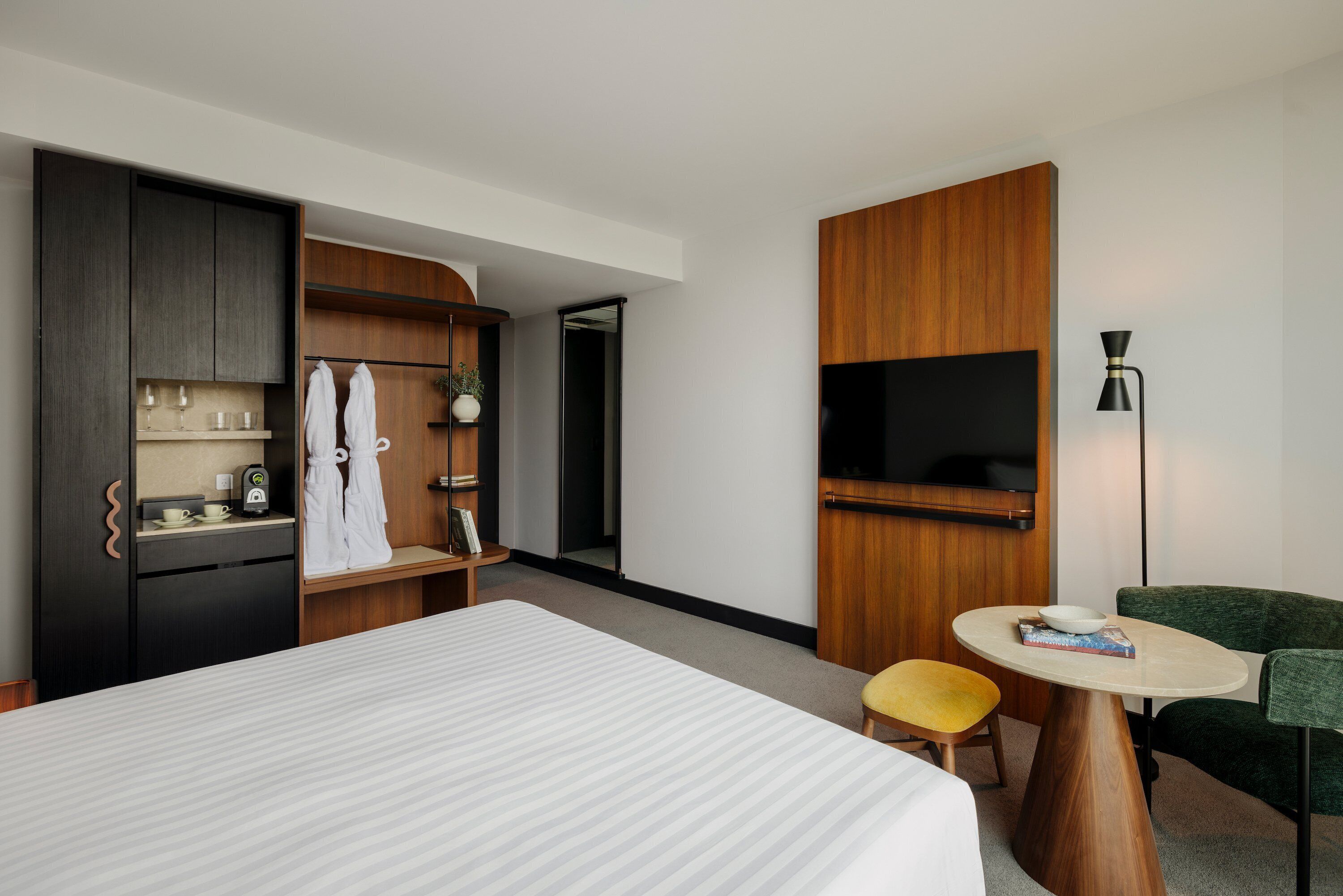 A hotel room with a bed, a small round table with a bowl and book, a green armchair, a wooden wardrobe, a TV mounted on the wall, and a small stool.