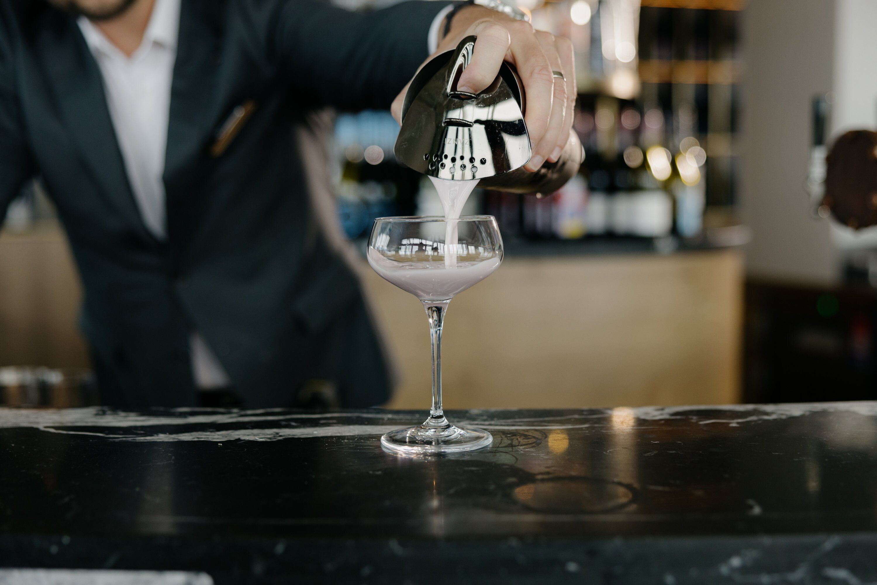 A bartender pouring a drink into a martini glass on a bar counter.
