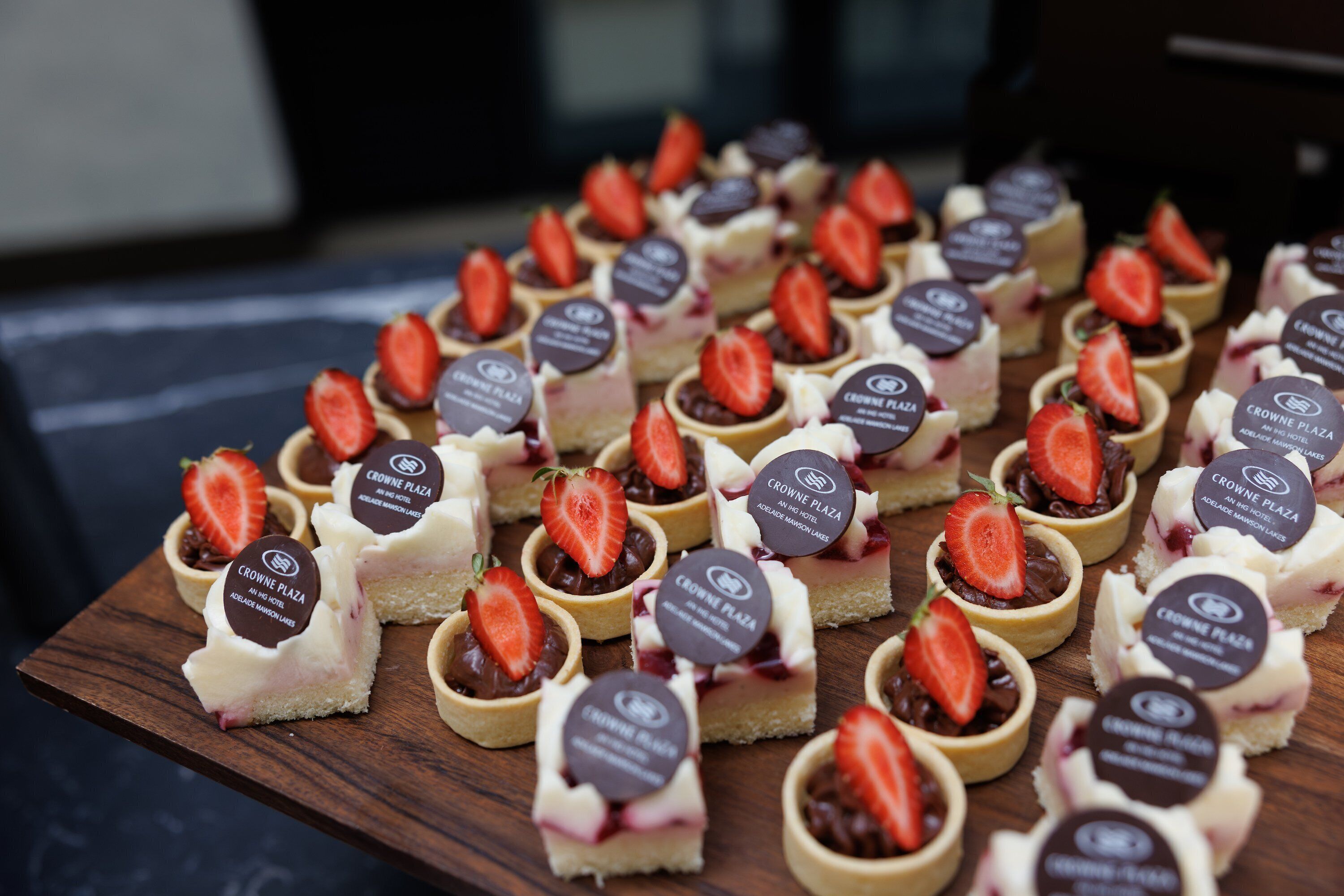 A wooden board with multiple small dessert items, each topped with a strawberry and labeled "Crowne Plaza."
