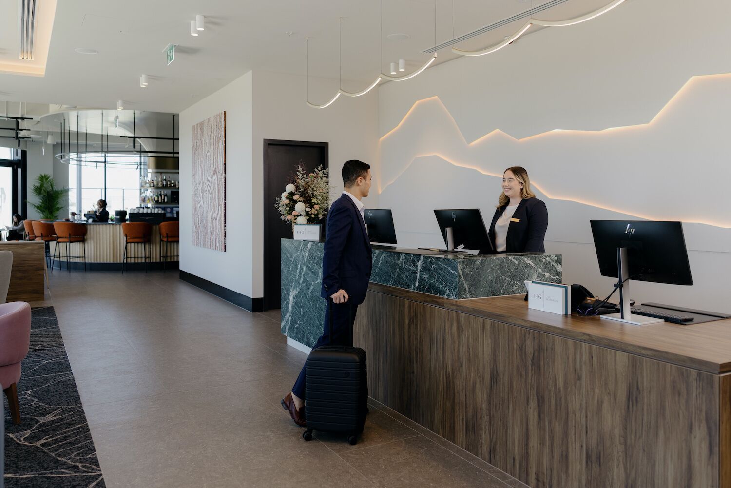 A modern hotel reception area with a marble counter, two receptionists, and a man with a suitcase.