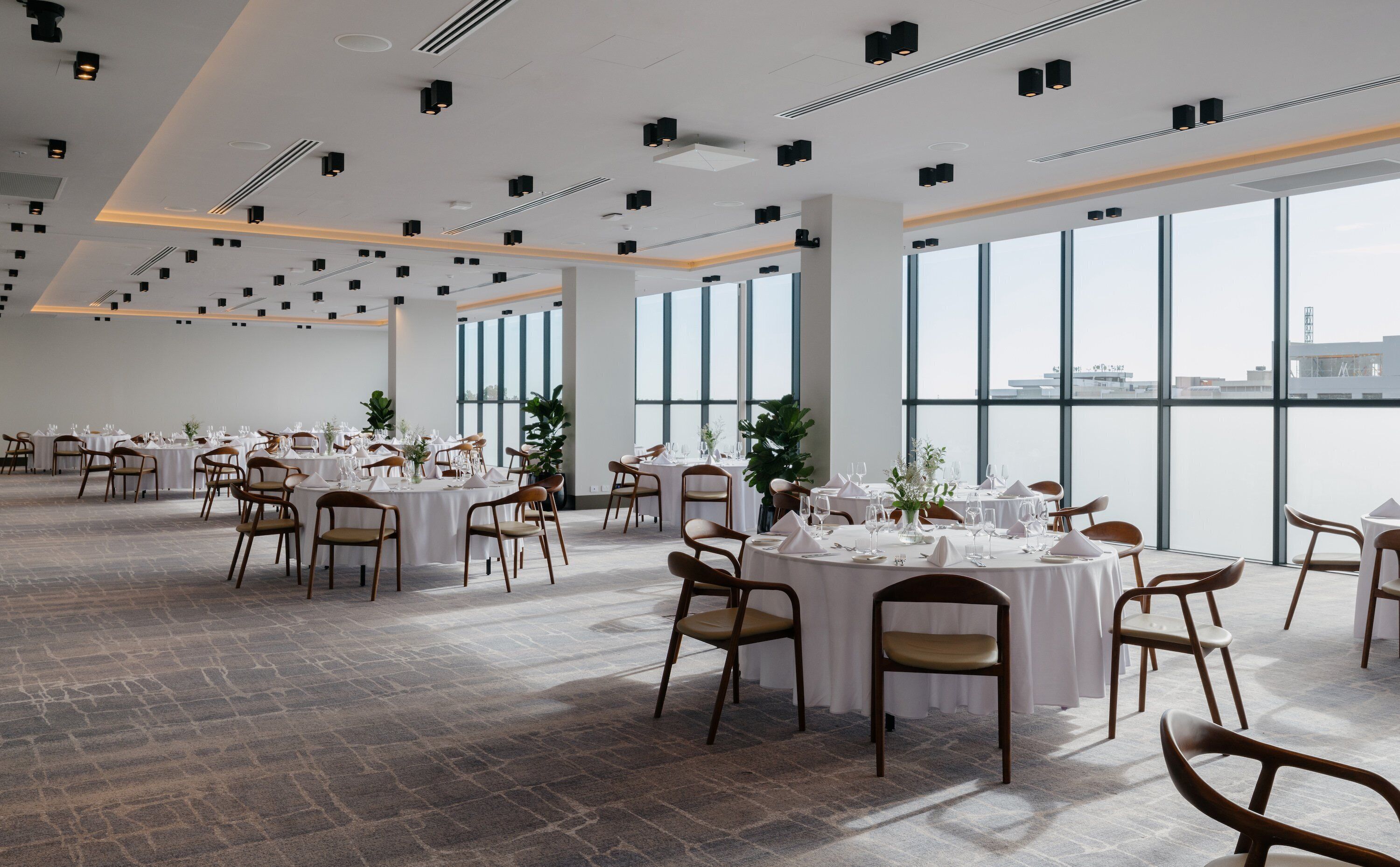 A spacious dining area with round tables and chairs, large windows offering a view of the city, and a modern ceiling design.