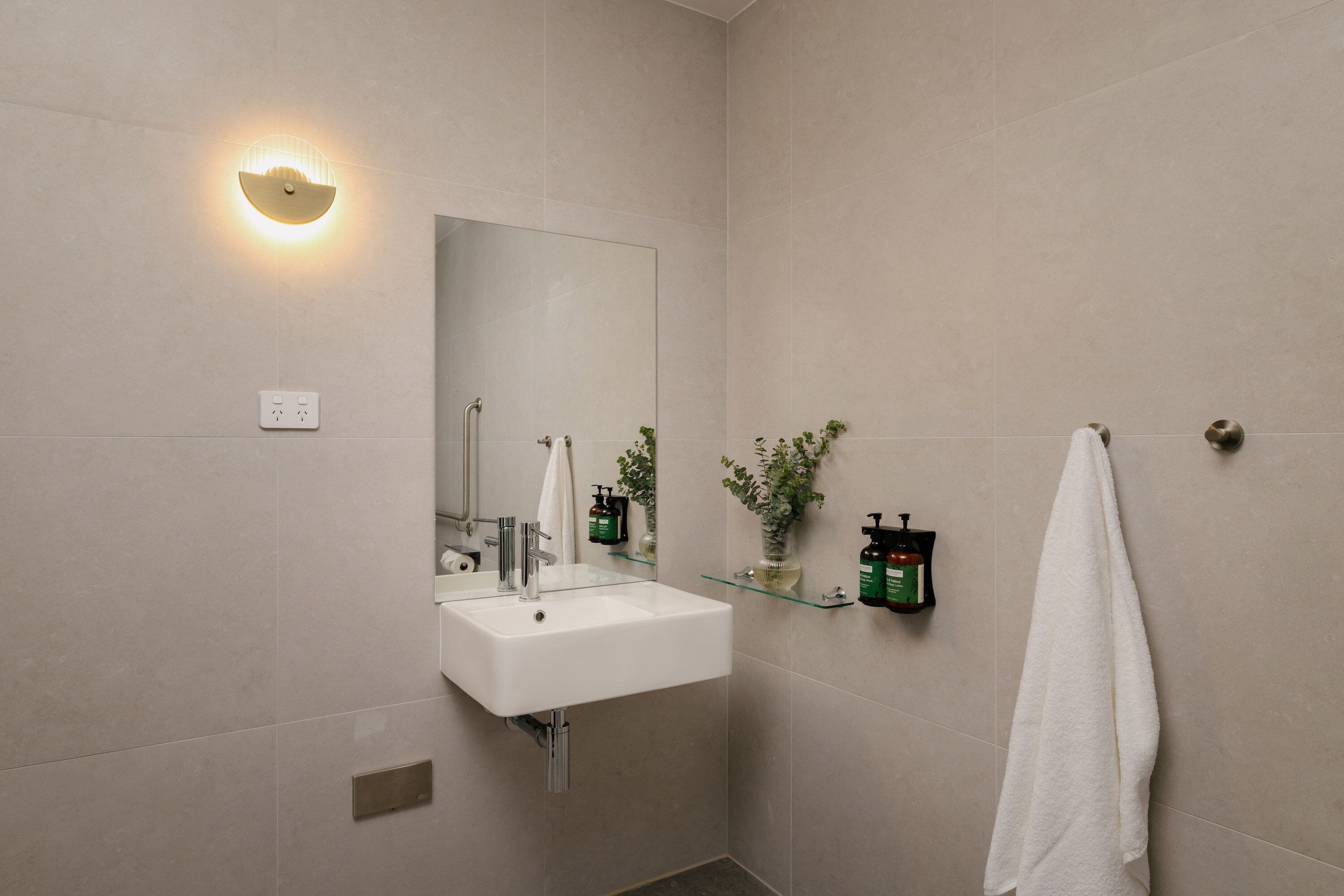 A modern bathroom with a white sink, a glass shelf with toiletries, a towel hanging on the wall, and a wall-mounted light.
