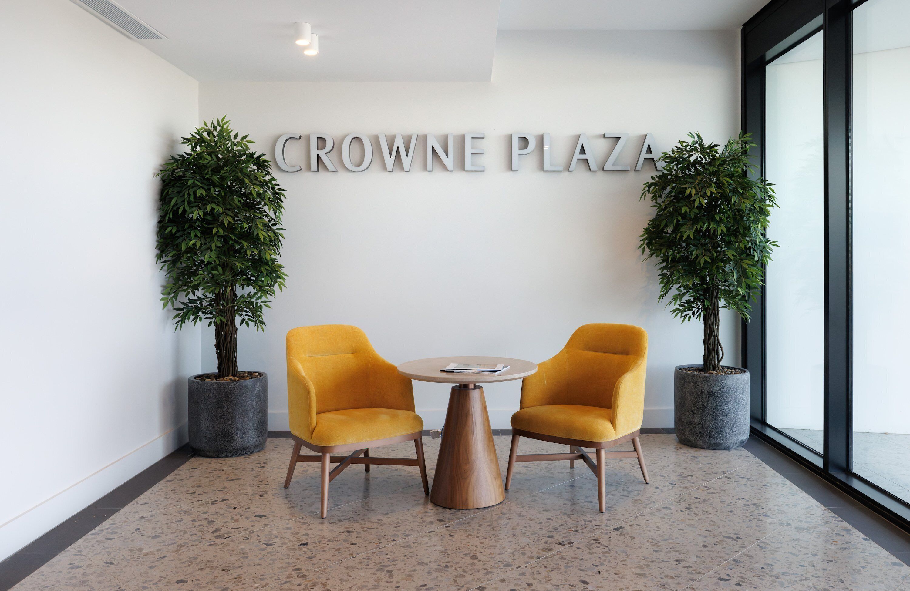 A modern interior with two yellow chairs around a round table, a sign reading "CROWNE PLAZA", and two potted plants.