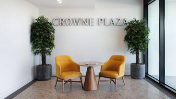 A modern interior with two yellow chairs around a round table, a sign reading "CROWNE PLAZA", and two potted plants.