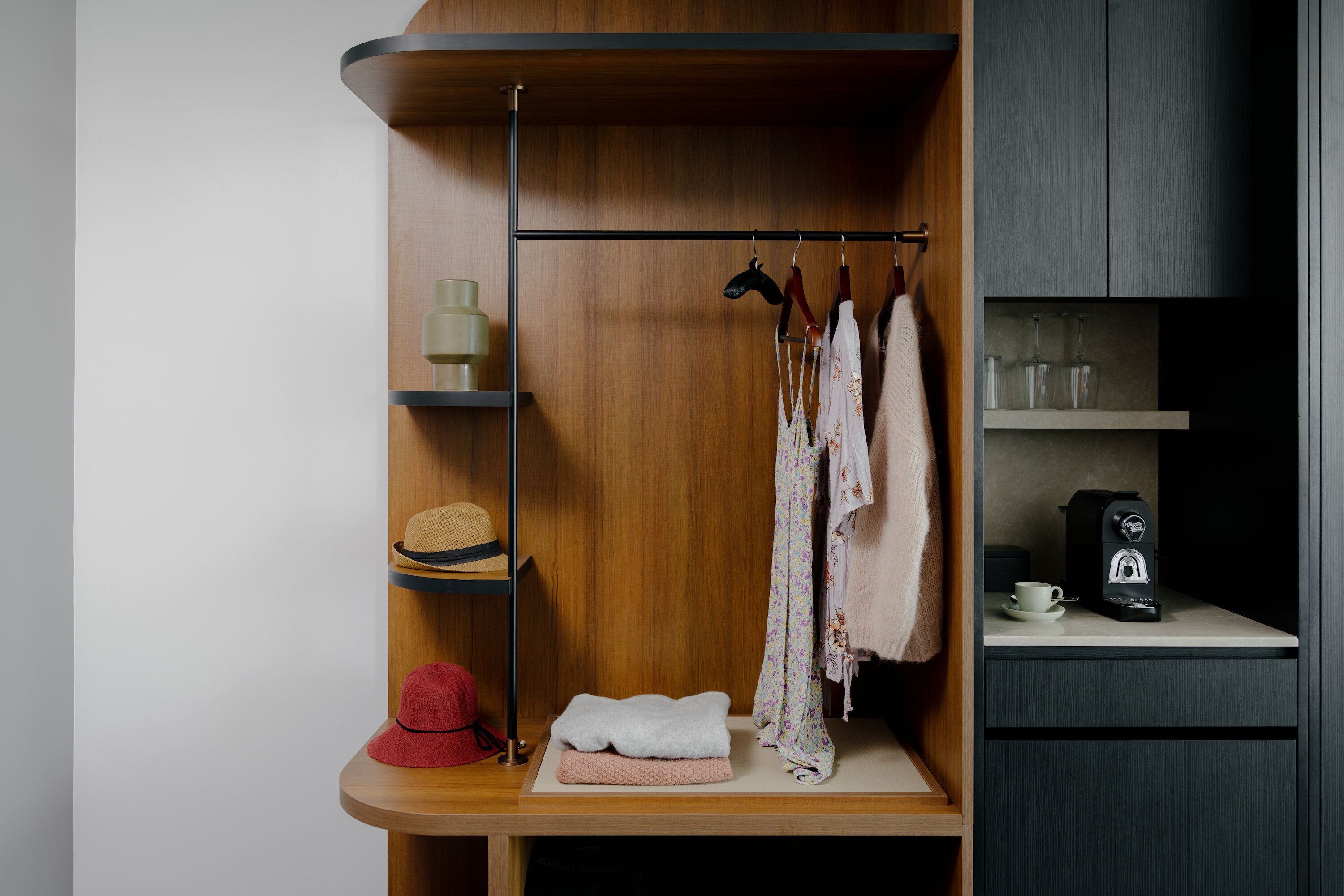 Een netjes geordende garderobe met hangende kleding, planken met hoeden en een koffieapparaat op een aanrecht.