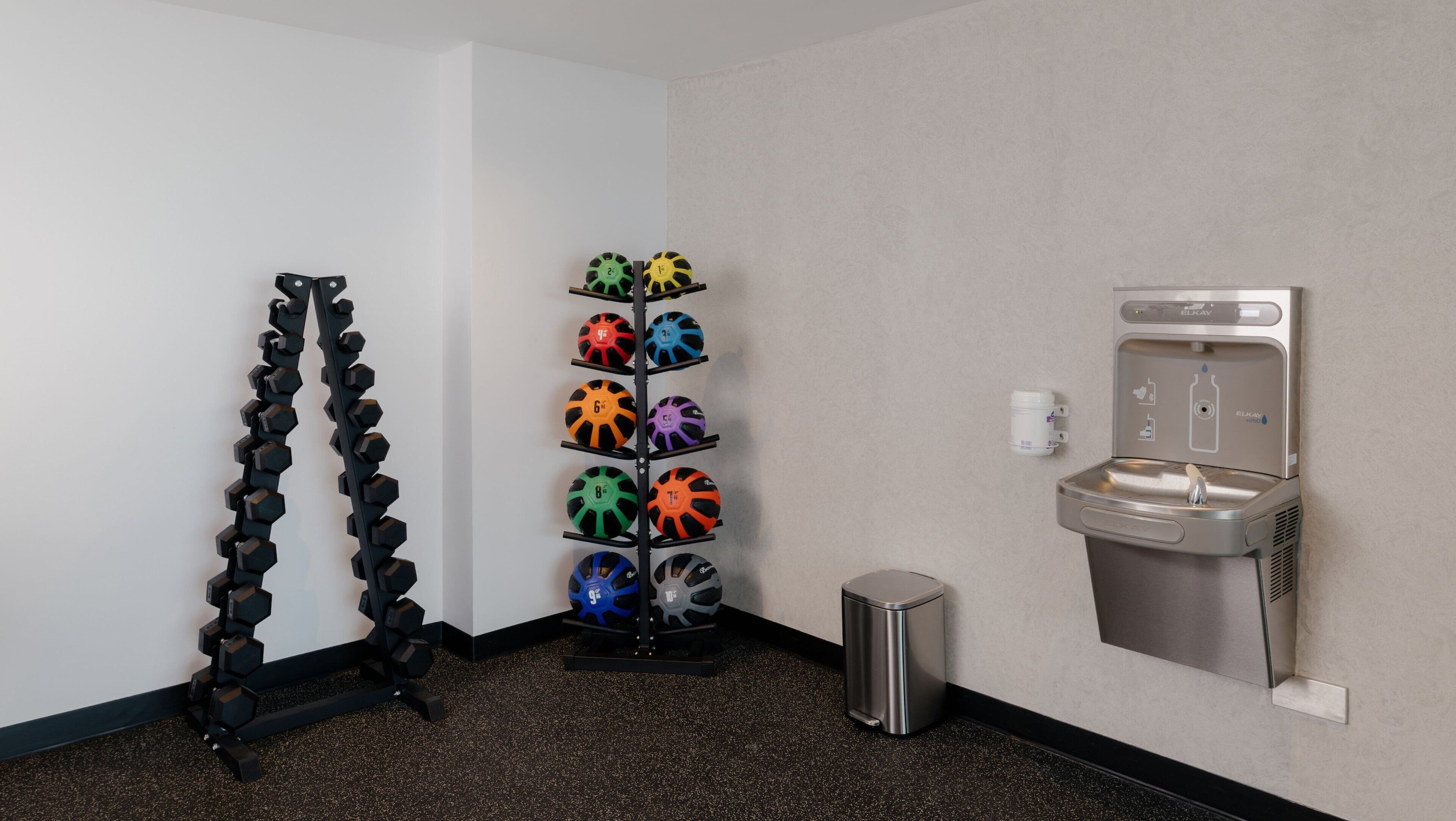A gym area with a rack of dumbbells, a wall-mounted water dispenser, and a trash bin.