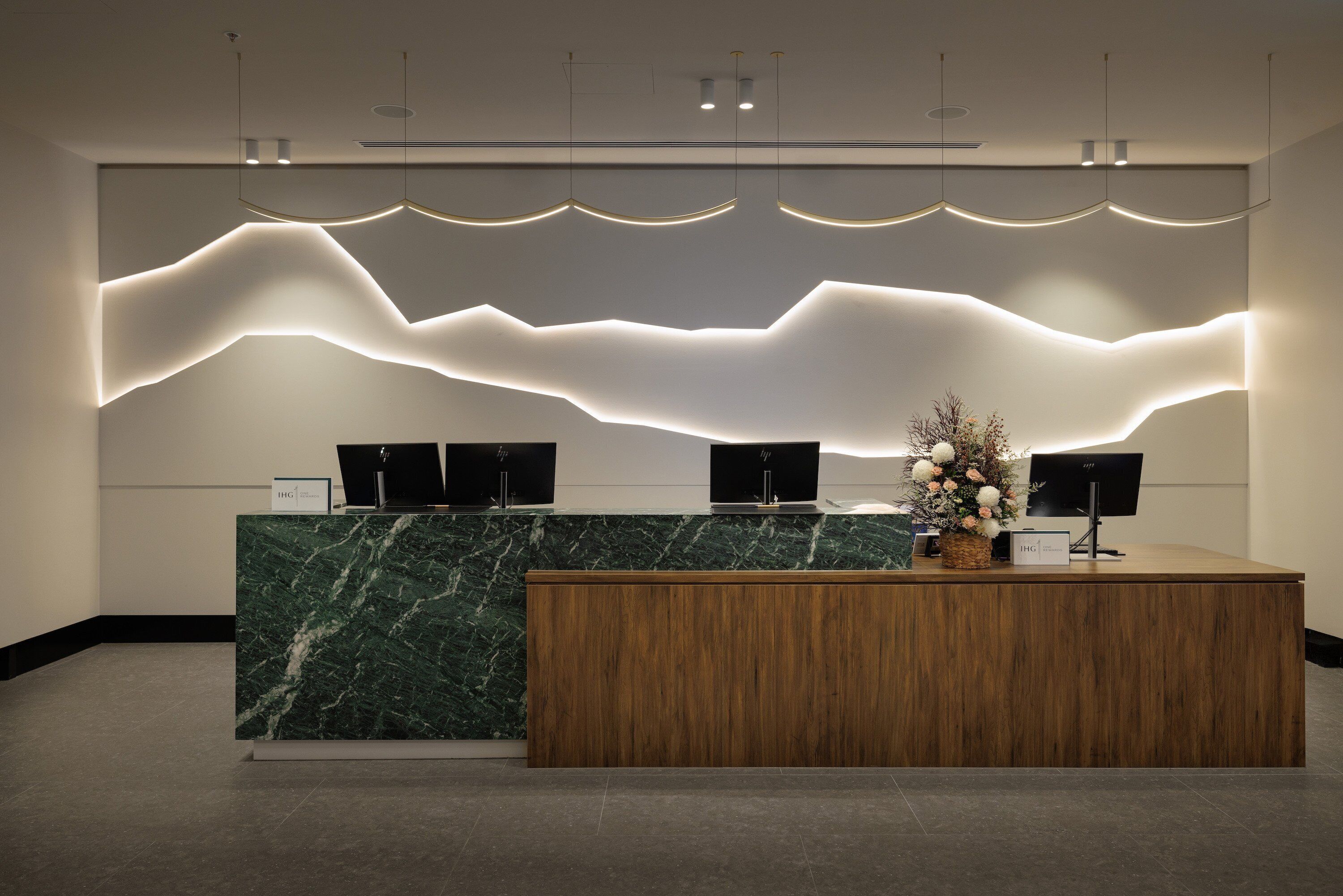A reception desk with a marble top and wooden base, flanked by two monitors and a decorative plant.