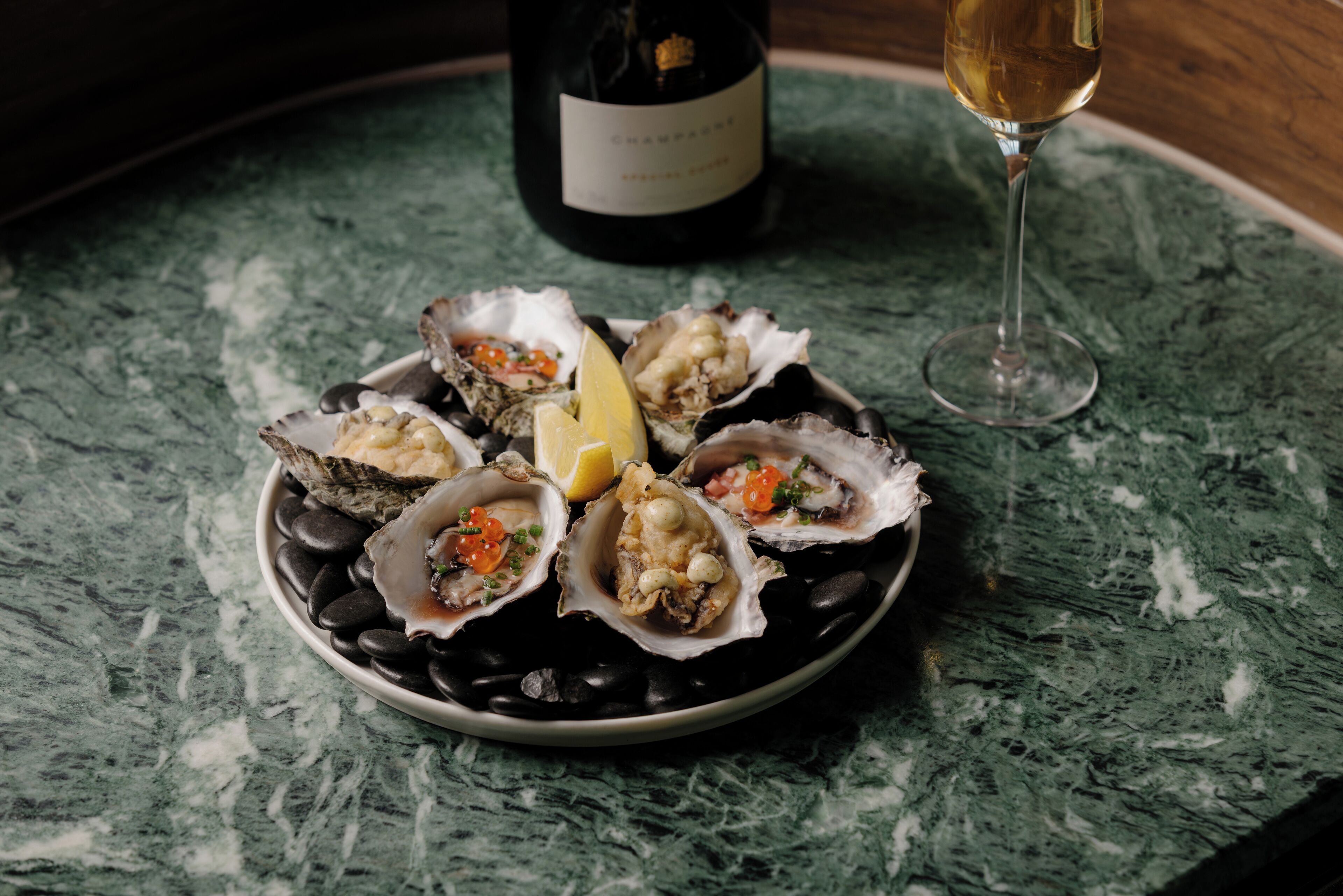 A plate of oysters with lemon and sauce, a bottle of champagne, and a glass of wine on a marble table.