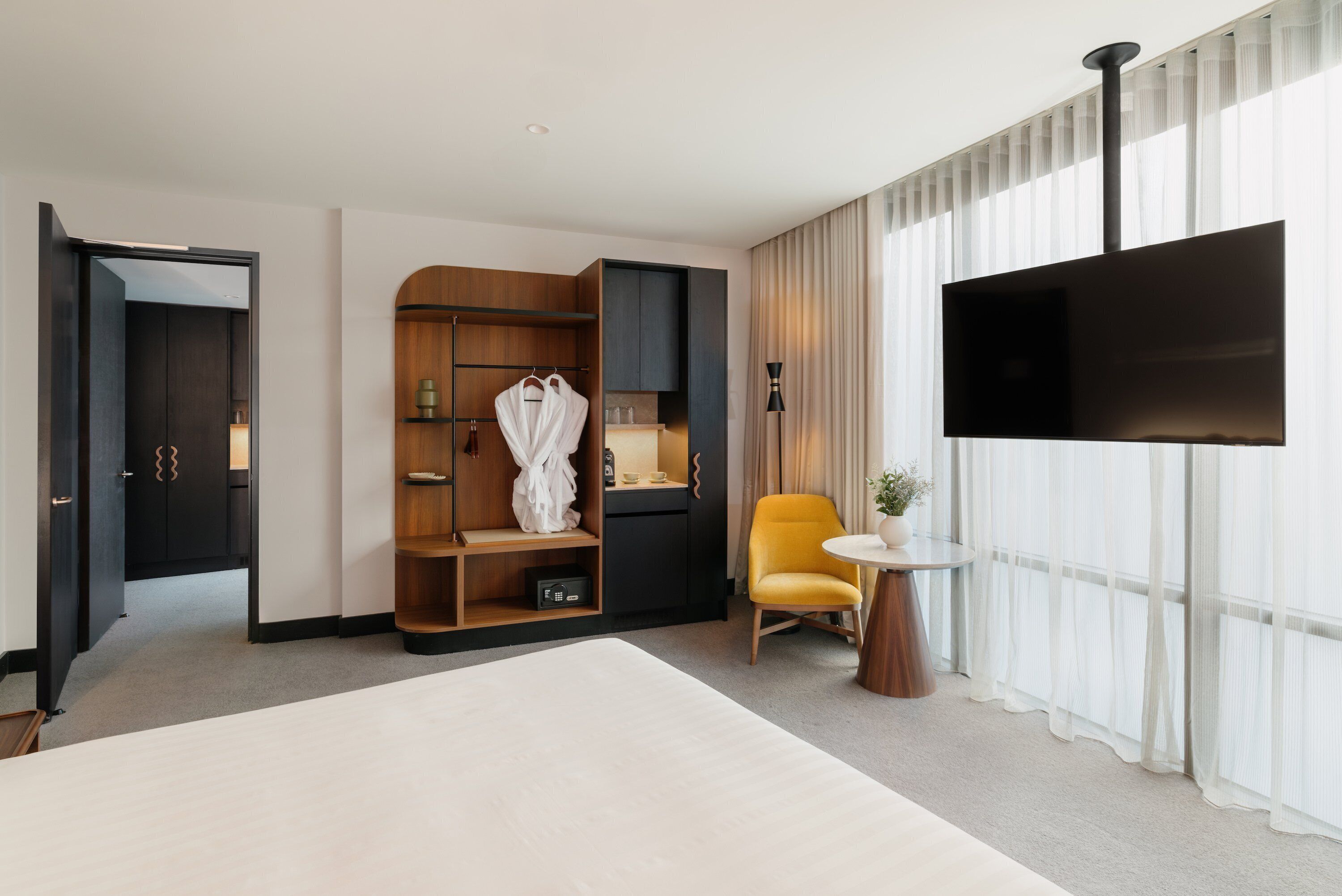 A hotel room with a large bed, a wardrobe with a mirror, a yellow armchair, a small round table with a plant, and a TV mounted on the wall.