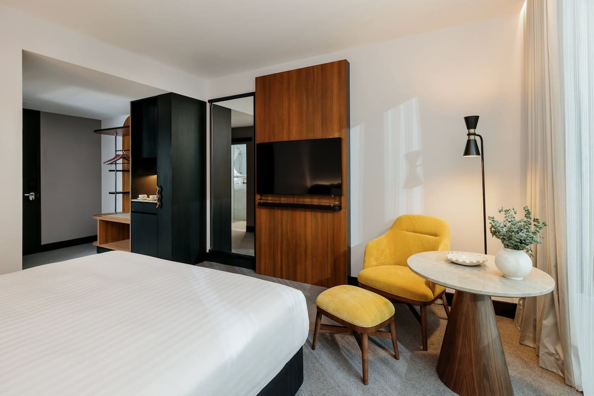 Ein Hotelzimmer mit einem großen Bett, einem gelben Sessel, einem kleinen runden Tisch mit einer Vase und einem Schrank mit Fernseher.
