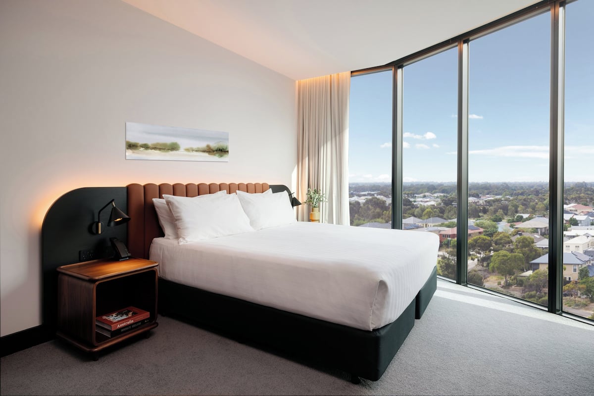 Ein modernes Hotelzimmer mit einem großen Bett, einem Nachttisch, Blick auf die Stadt durch raumhohe Fenster und einem Landschaftsbild an der Wand.