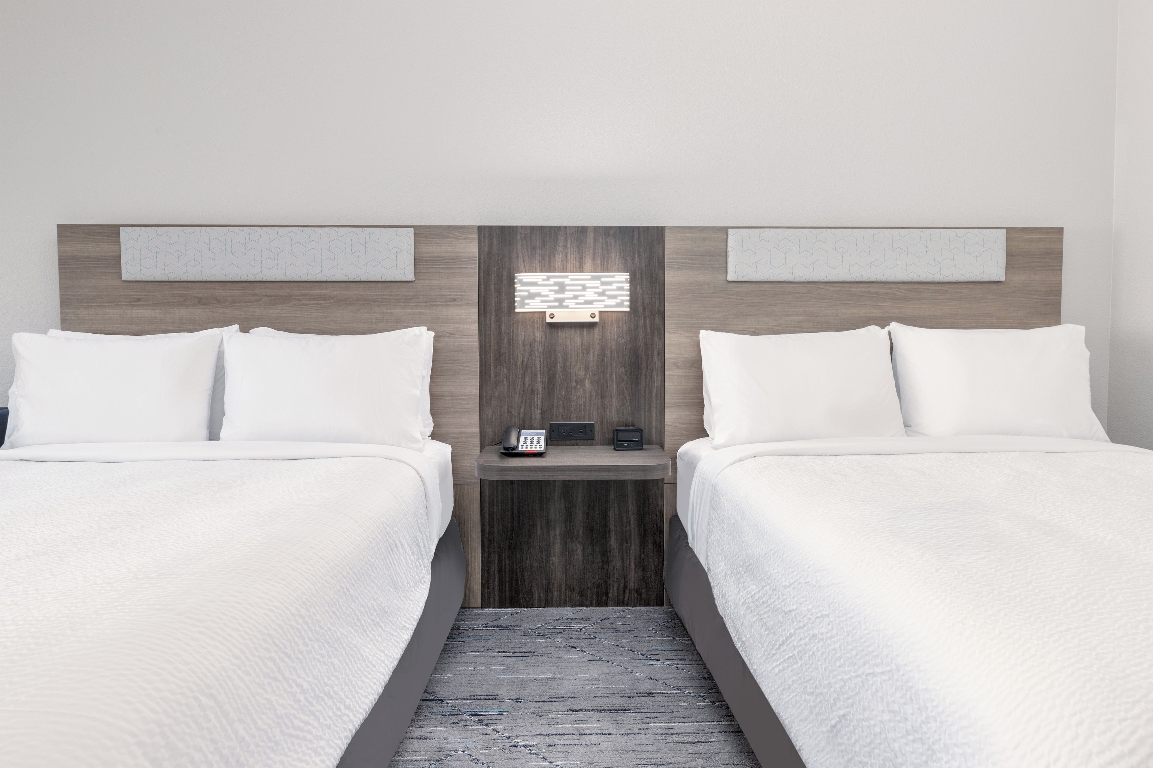 A hotel room with two beds, a bedside table with a phone and a small clock, and a wall-mounted light fixture.