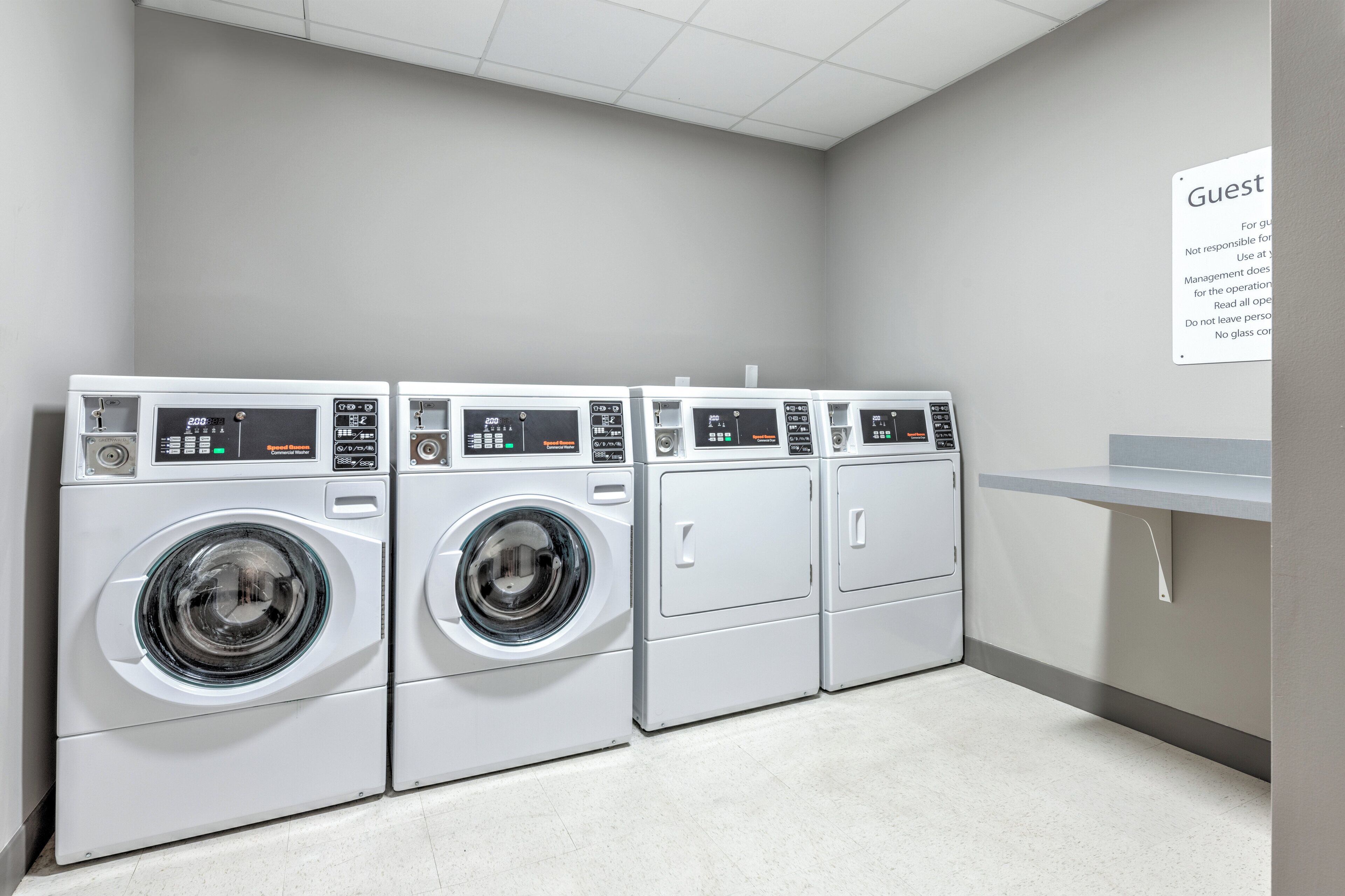 A row of four white front-loading washing machines in a laundry room with a sign on the wall and a small counter to the right.